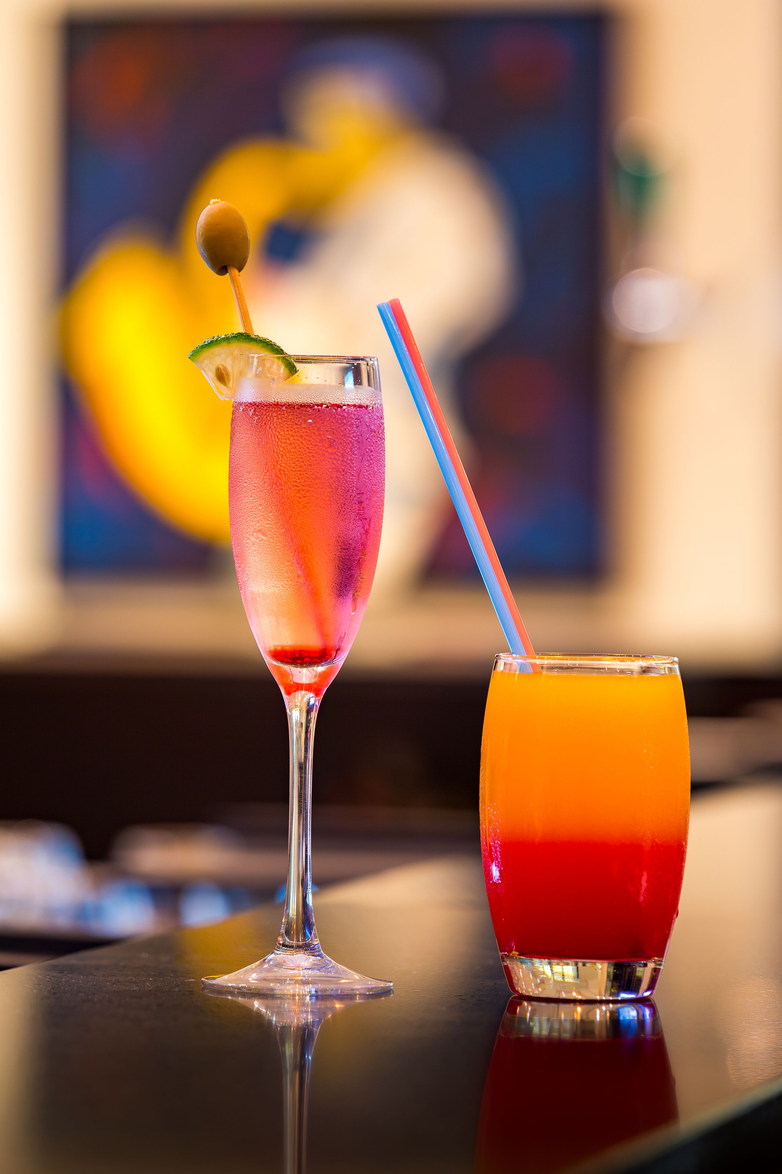 Two glasses of drinks on a table with a blurred painting in the background.