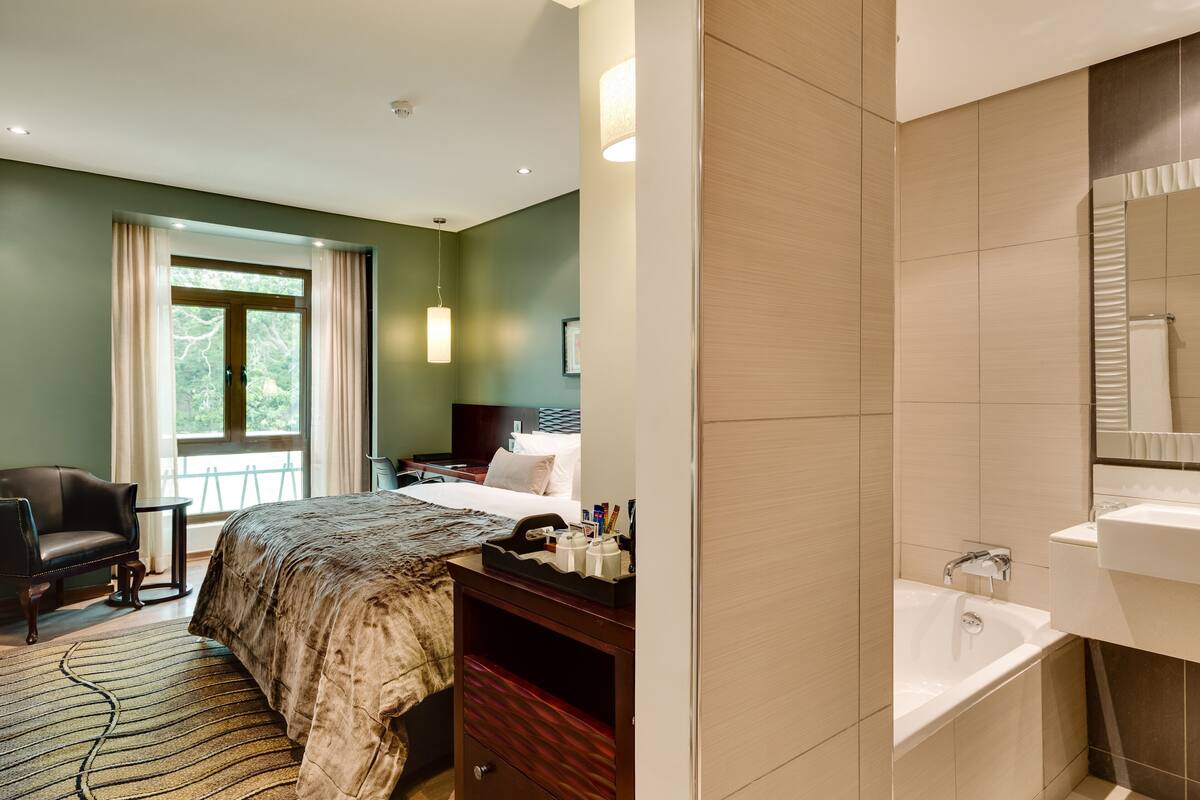 Habitación de hotel con cama, escritorio, silla, ventana con cortinas y baño con lavamanos y espejo.
