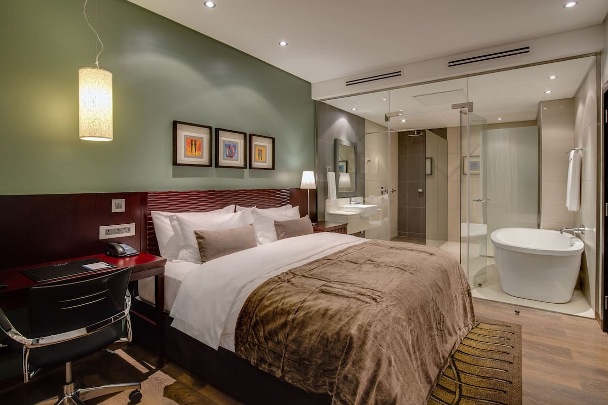 Habitación de hotel con cama, escritorio, silla, mesita de noche, lámpara y baño con ducha, lavabo y bañera.