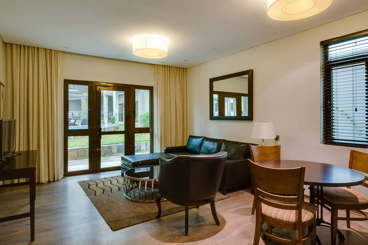 Un salón moderno con sofá, una mesa de comedor y sillas. Cuenta con un ventanal con persianas, un espejo en la pared y una lámpara sobre la mesa.