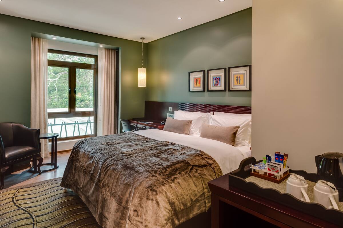 Habitación de hotel con una cama grande, un escritorio con cafetera, una silla y vistas al exterior a través de un ventanal.