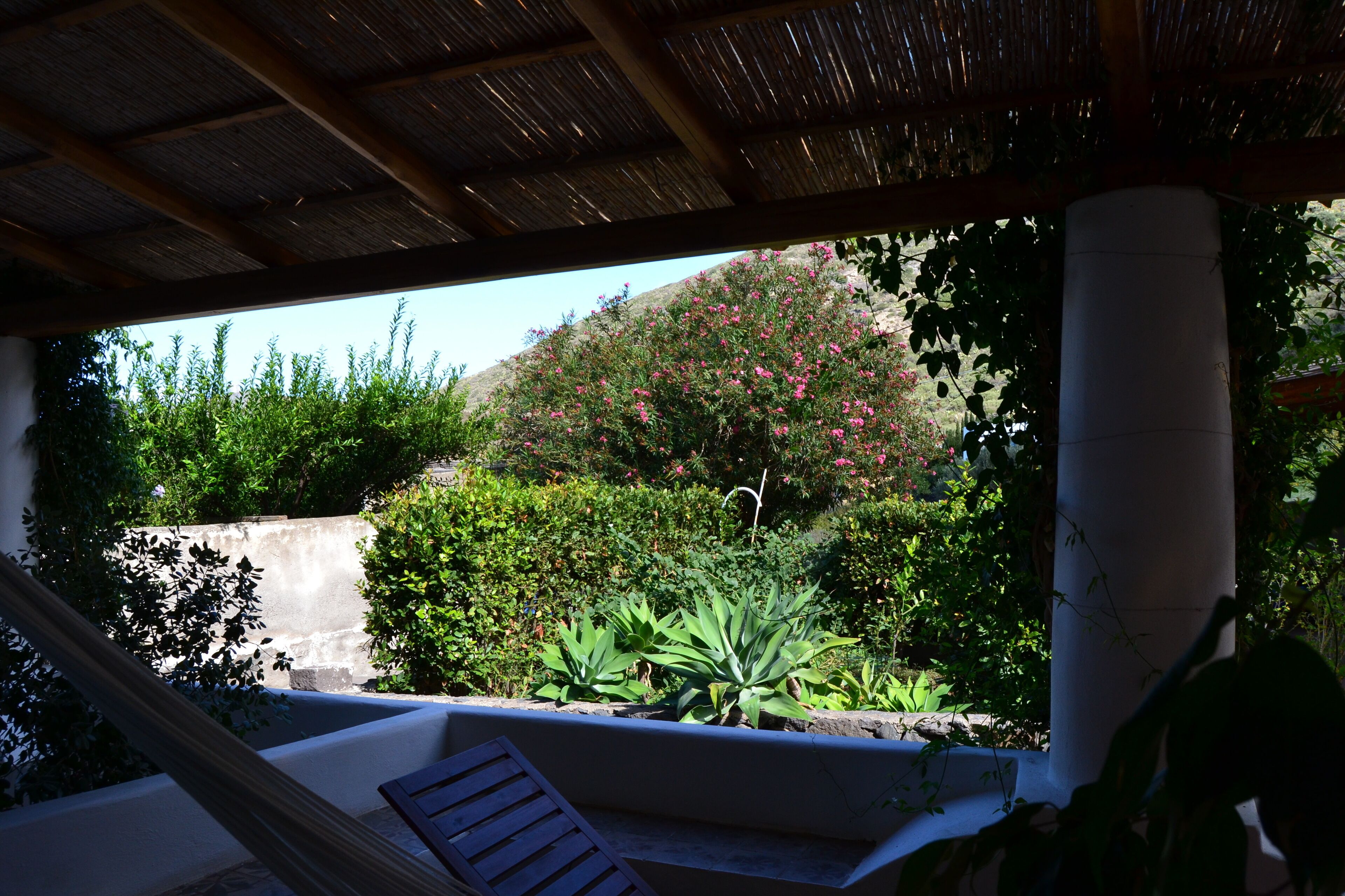 A covered outdoor seating area with a view of a garden and a hill.