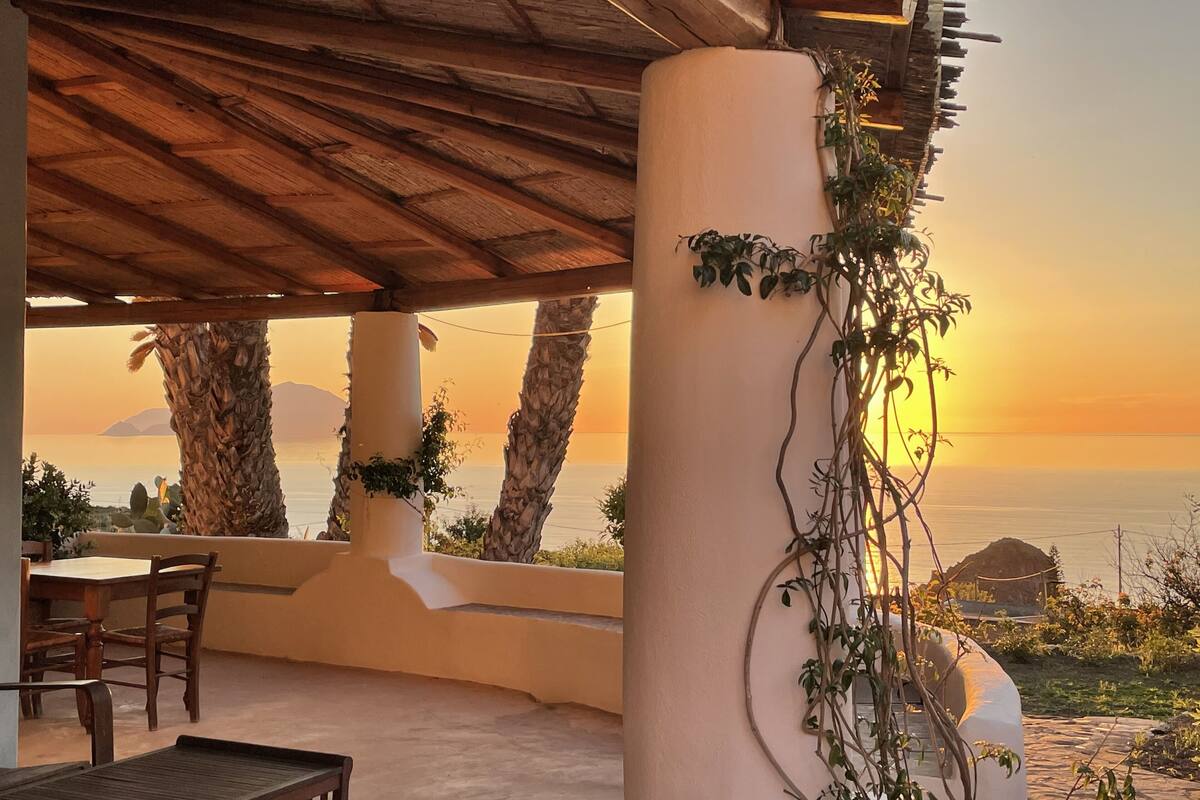 A covered outdoor seating area with a view of the sunset over the sea, wooden furniture, and a column with climbing plants.