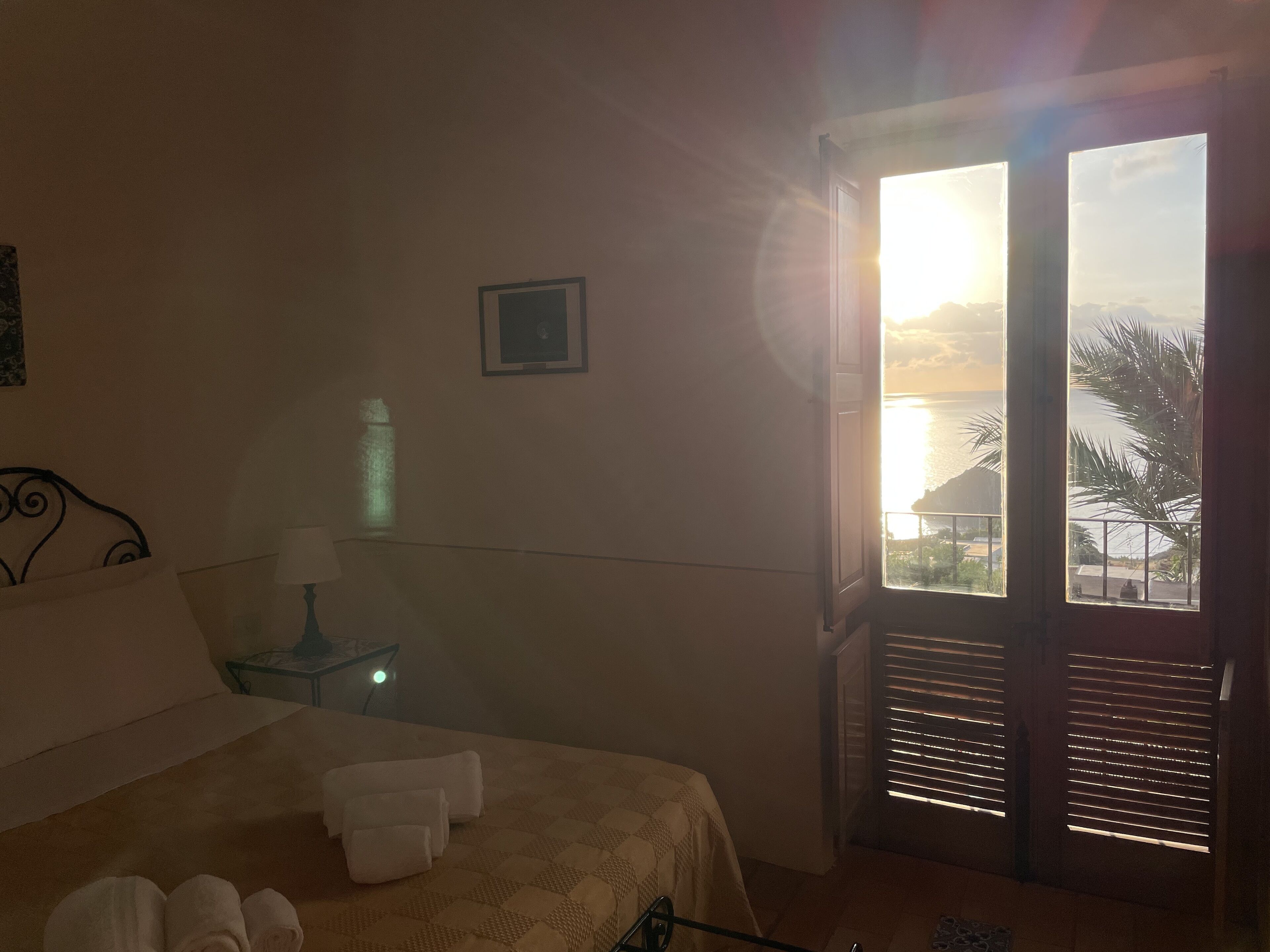 A hotel room with a bed, a nightstand, a lamp, a framed picture on the wall, and a view of the sea through the window.
