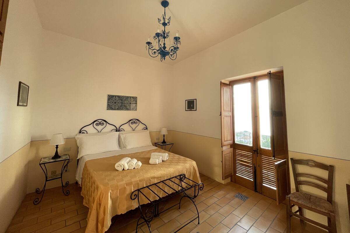 A bedroom with a bed, two bedside tables, a chandelier, a wooden door, and a chair.