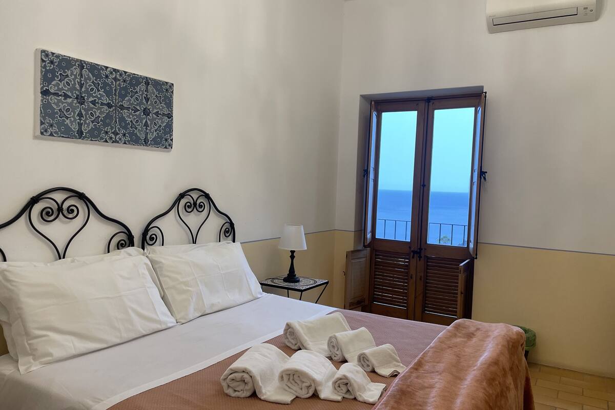A neatly made bed with white linens and a brown blanket, a bedside table with a lamp, and a window with a view of the sea.