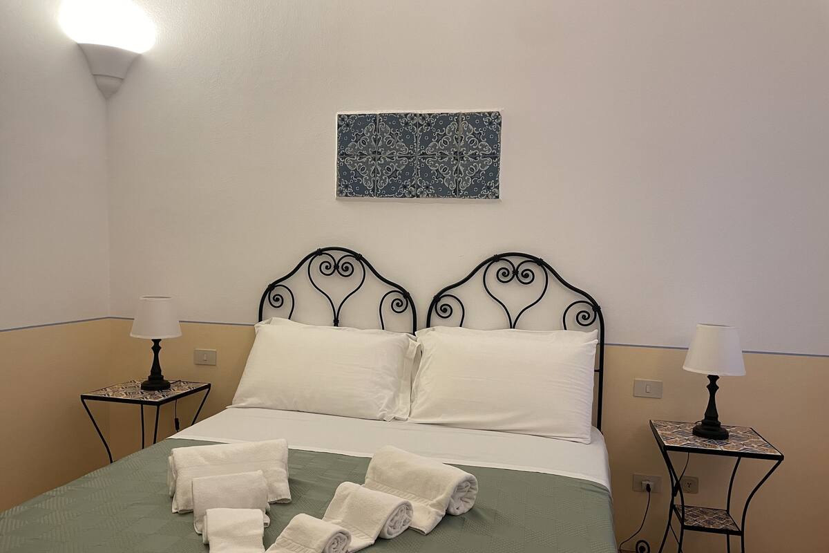 A neatly made bed with white linens and rolled towels, two bedside tables with lamps, and a decorative wall piece above the bed.