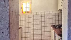 Ett smalt kök med kakel på väggen bakom arbetsbänken, ett fönster med gula karmar och ett tegelgolv.