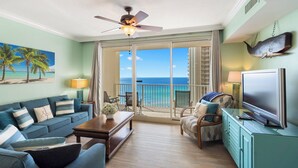 A spacious living room with a large sofa, a coffee table, and a TV. A balcony with a view of the ocean is visible. - 2BR/2BA, Amazing Gulf/Pool Views! Shores of Panama (Panama City Beach)
