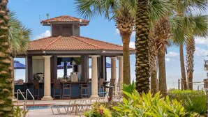 A poolside area with a covered bar, lounge chairs, and palm trees. - 2BR/2BA, Amazing Gulf/Pool Views! Shores of Panama (Panama City Beach)