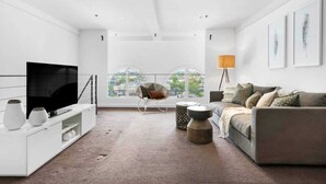 A modern living room with a large flat-screen TV, a grey sofa, a white entertainment unit, and a large window with a view of the city. - Light-filled Converted Warehouse 2 Bedroom Apartment in Prahran (Prahran)