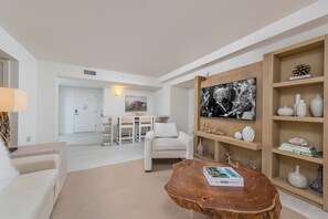 Uma sala de estar moderna com uma mesa de centro de madeira, um sofá branco, uma área de jantar com mesa e cadeiras brancas e uma estante com objetos decorativos.