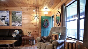 A cozy cabin interior with a plush armchair, a wooden coffee table, a patterned rug, and a green door adorned with a wreath. - Private Mountain Cabin to Gather, Relax & Unwind near Pigeon Forge,TN (Sevierville)