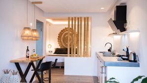 A modern kitchen with a central island, a dining area with a table and chairs, and a bedroom with a bed and decorative wall. - Case di Venia (Mykonos)