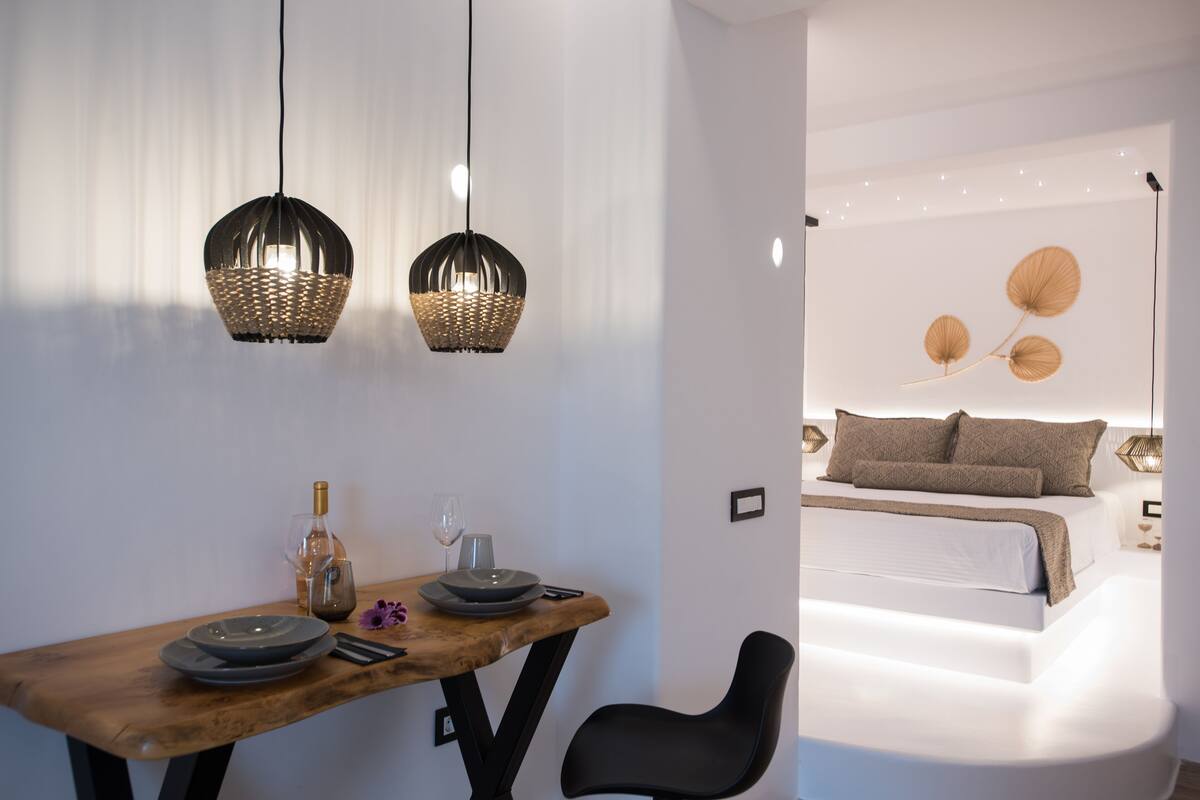 Una habitación moderna con una mesa de comedor de madera, dos lámparas colgantes de mimbre, una cama con ropa de cama blanca y un aplique de pared.