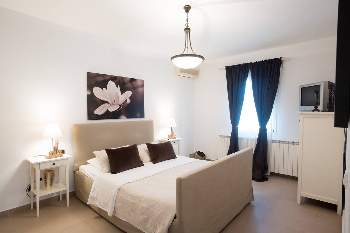 Un dormitorio con cama, mesitas de noche, lámpara, televisor y ventana con cortinas.