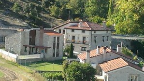 Un edificio de dos plantas con fachada de piedra y techo de tejas rojas, rodeado de vegetación y una ladera.