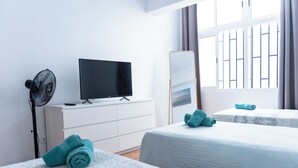 Quarto de hotel com duas camas, uma televisão de tela plana, um ventilador e uma janela grande com cortinas.