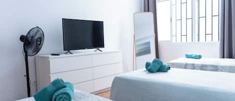 A hotel room with two beds, a flat-screen TV, a standing fan, and a large window with curtains.