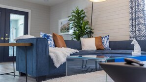 A modern living room with a dark blue sofa, white throw pillows, a glass coffee table, and a potted plant. - Coffee Lovers Paradise (Scottsdale)