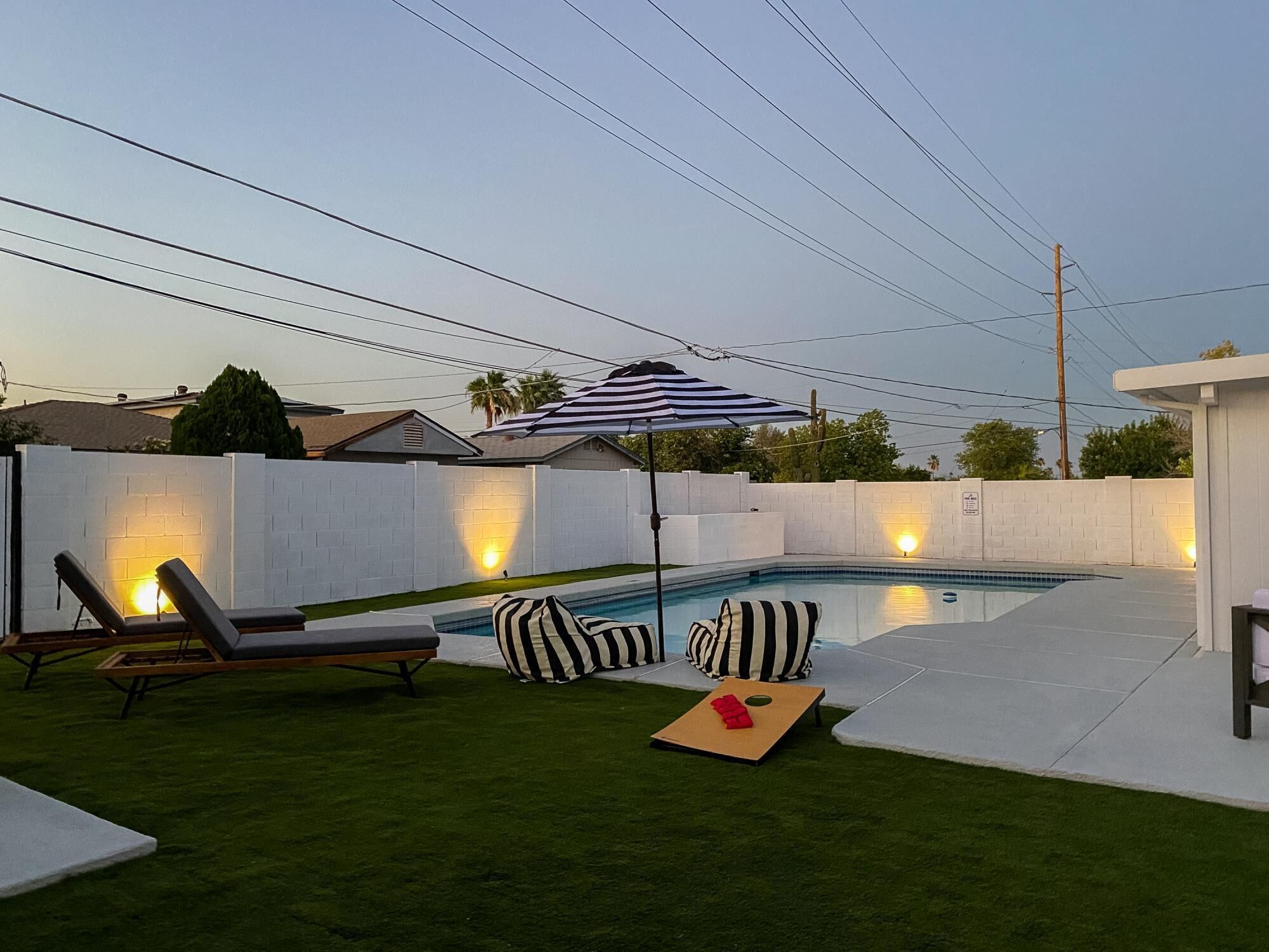 A backyard with a swimming pool, lounge chairs, a striped umbrella, and artificial grass.