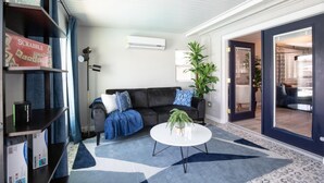 A living room with a black sofa, a white coffee table, and a blue patterned rug. There are shelves with board games and a potted plant. - Coffee Lovers Paradise (Scottsdale)
