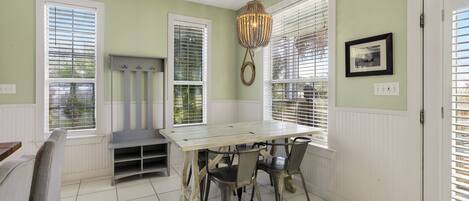A dining area with a wooden table and chairs, a hanging light fixture, and windows with blinds.