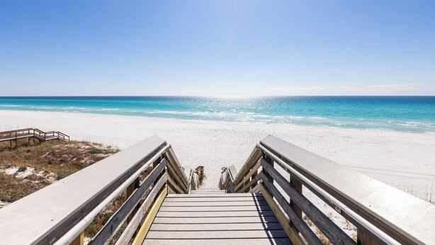 Une promenade en bois qui mène à une plage où l’eau est d’un bleu limpide et le rivage est sablonneux.