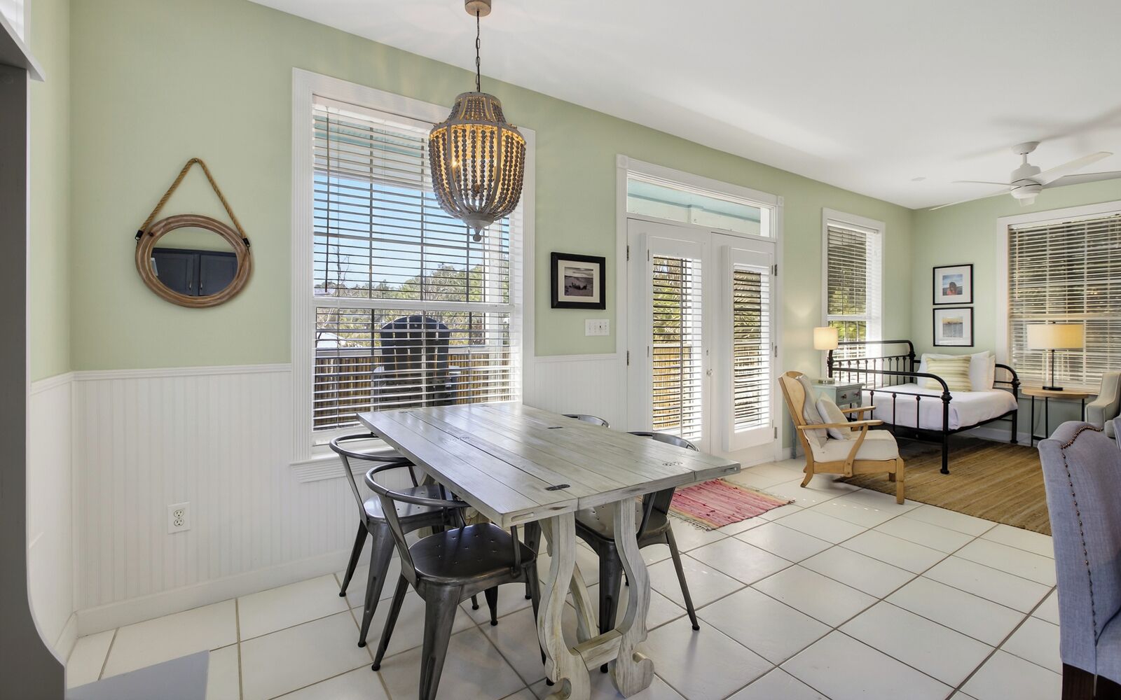 A dining area with a wooden table and chairs, a hanging light fixture, and a view through large windows with blinds.