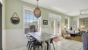 Un espace repas comprenant une table et des chaises en bois, un luminaire suspendu et une vue dégagée sur l’extérieur grâce à de grandes fenêtres équipées de stores.