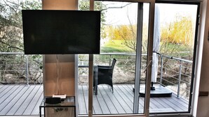 Ein modernes Wohnzimmer mit großem Fernseher, einem Glastisch und Blick auf eine Terrasse mit Gartenmöbeln.