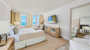 A hotel room with a large bed, a TV, a desk with a lamp, and a view of the sea.
