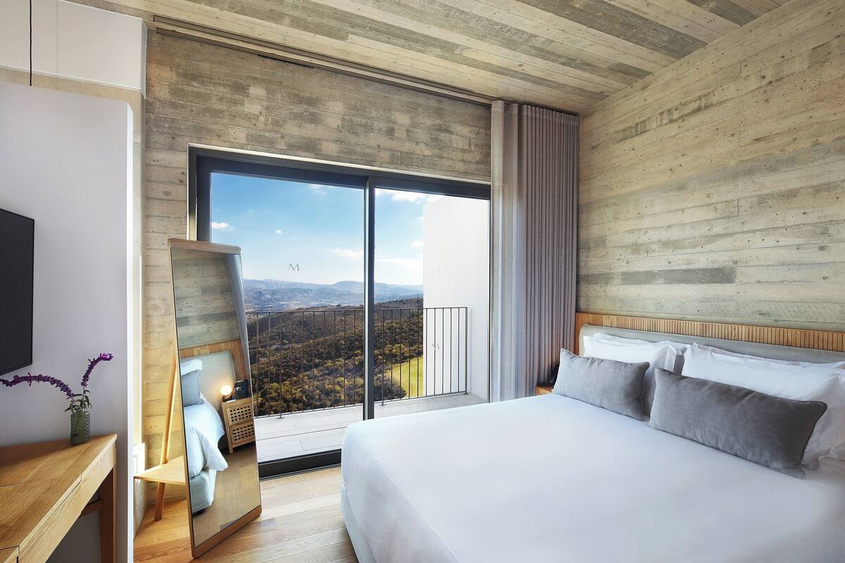 A modern hotel room with a large bed, a wooden headboard, a bedside table with a lamp, and a large window offering a scenic view.