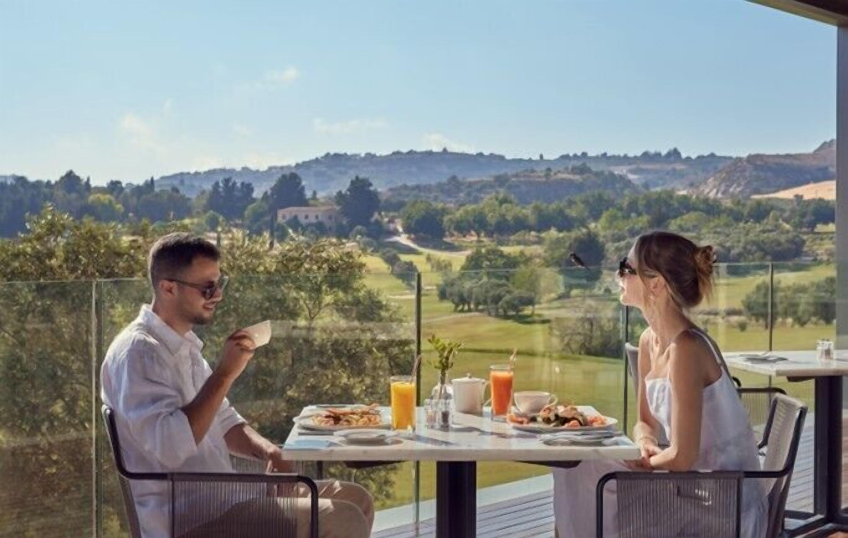 Una mesa de comedor con vistas a un campo de golf, dos personas sentadas y una vajilla con platos, vasos y un jarrón.