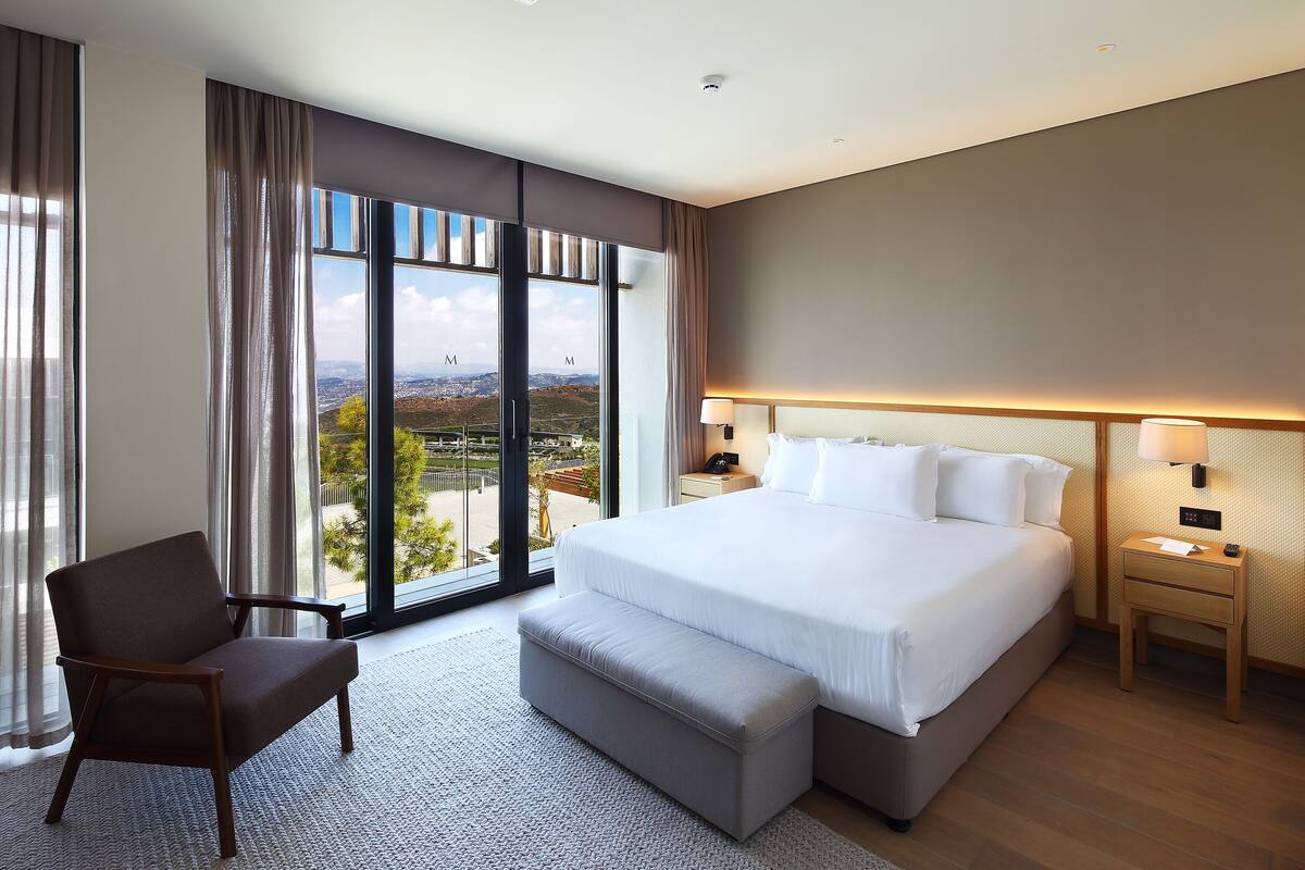 A hotel room with a large bed, a chair, a bench, a nightstand, and a view of the outdoors.