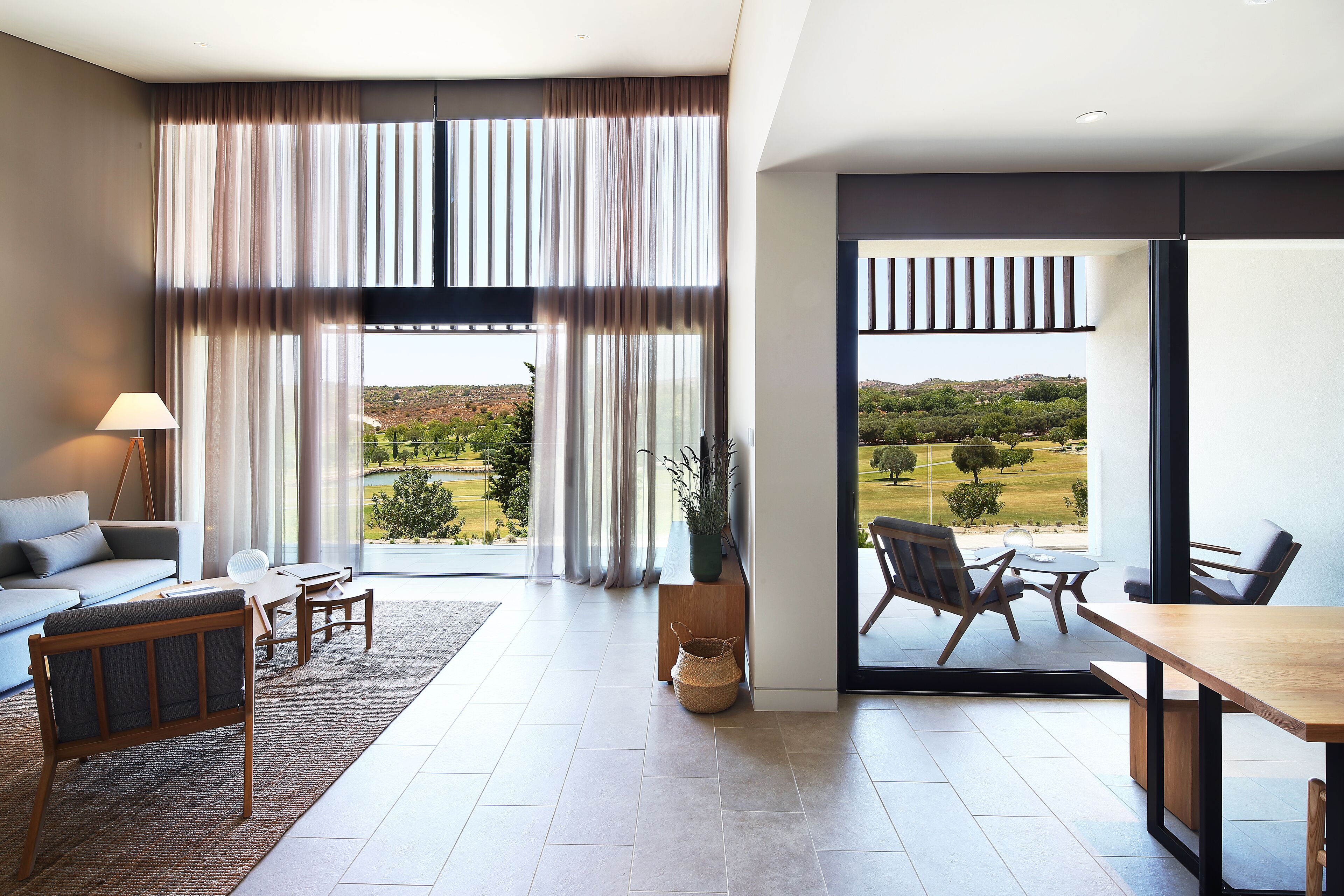 Ein modernes Wohnzimmer mit Sofa, Couchtisch und Esstisch. Ein großes Fenster mit Vorhängen, Blick auf eine Landschaft und ein Balkon mit Sitzgelegenheit.