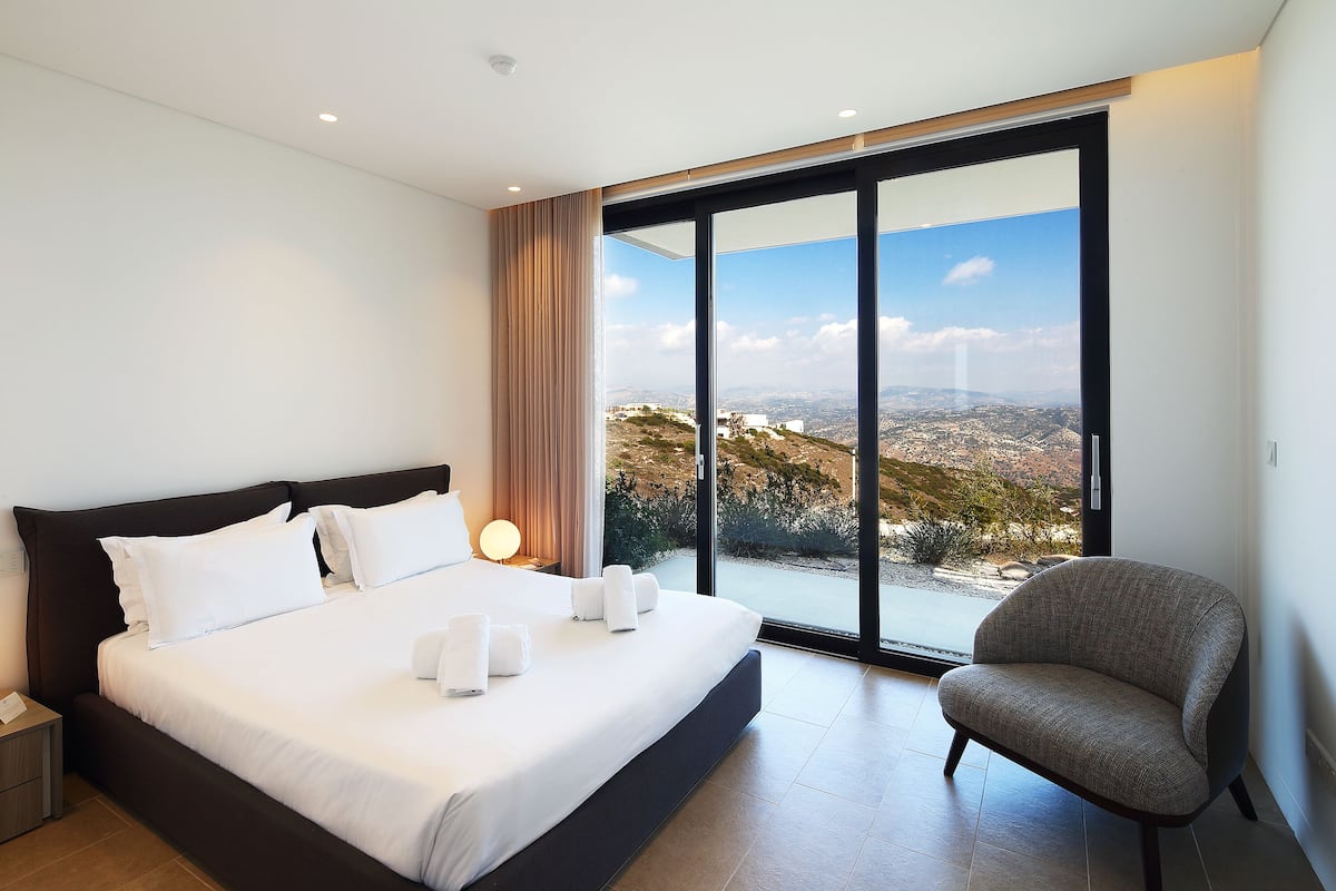 A modern bedroom with a large bed, a chair, and a view of a mountainous landscape through a sliding door.