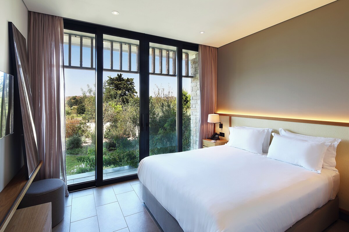 A modern hotel room with a large bed, a TV, and a view of greenery through the windows.