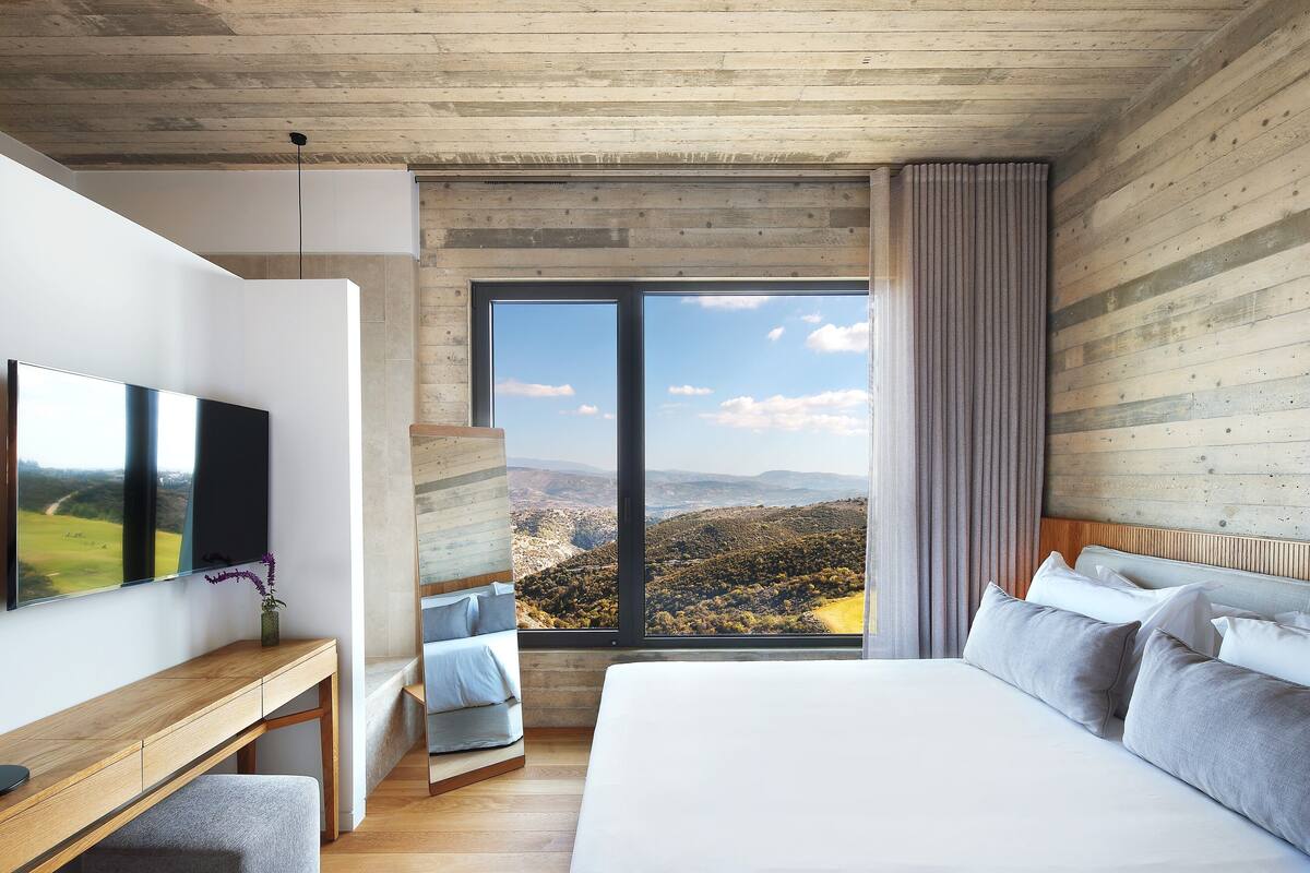 A modern bedroom with a large bed, a wooden desk, a flat-screen TV, and a large window offering a scenic view.