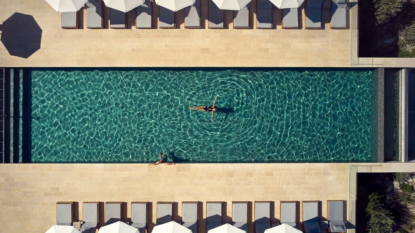 An aerial view of a rectangular swimming pool with clear water, surrounded by lounge chairs and umbrellas.