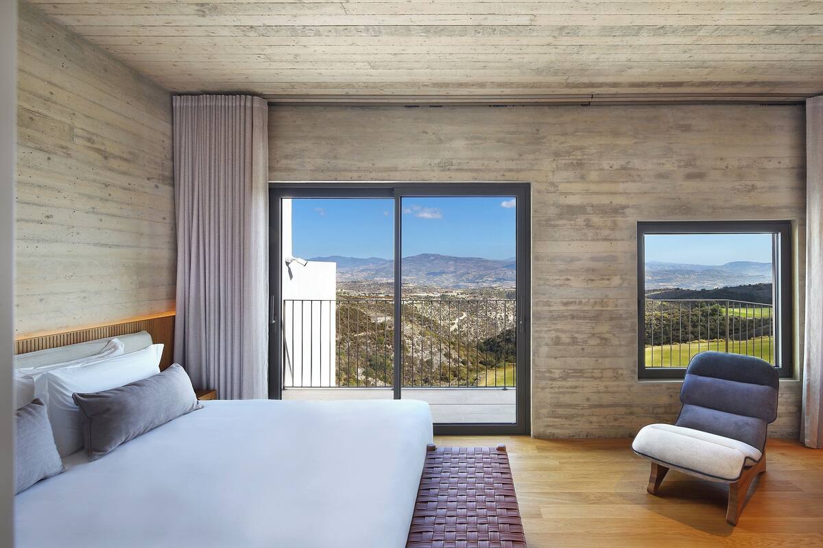 A bedroom with a large bed, a chair, and a balcony with a city view.