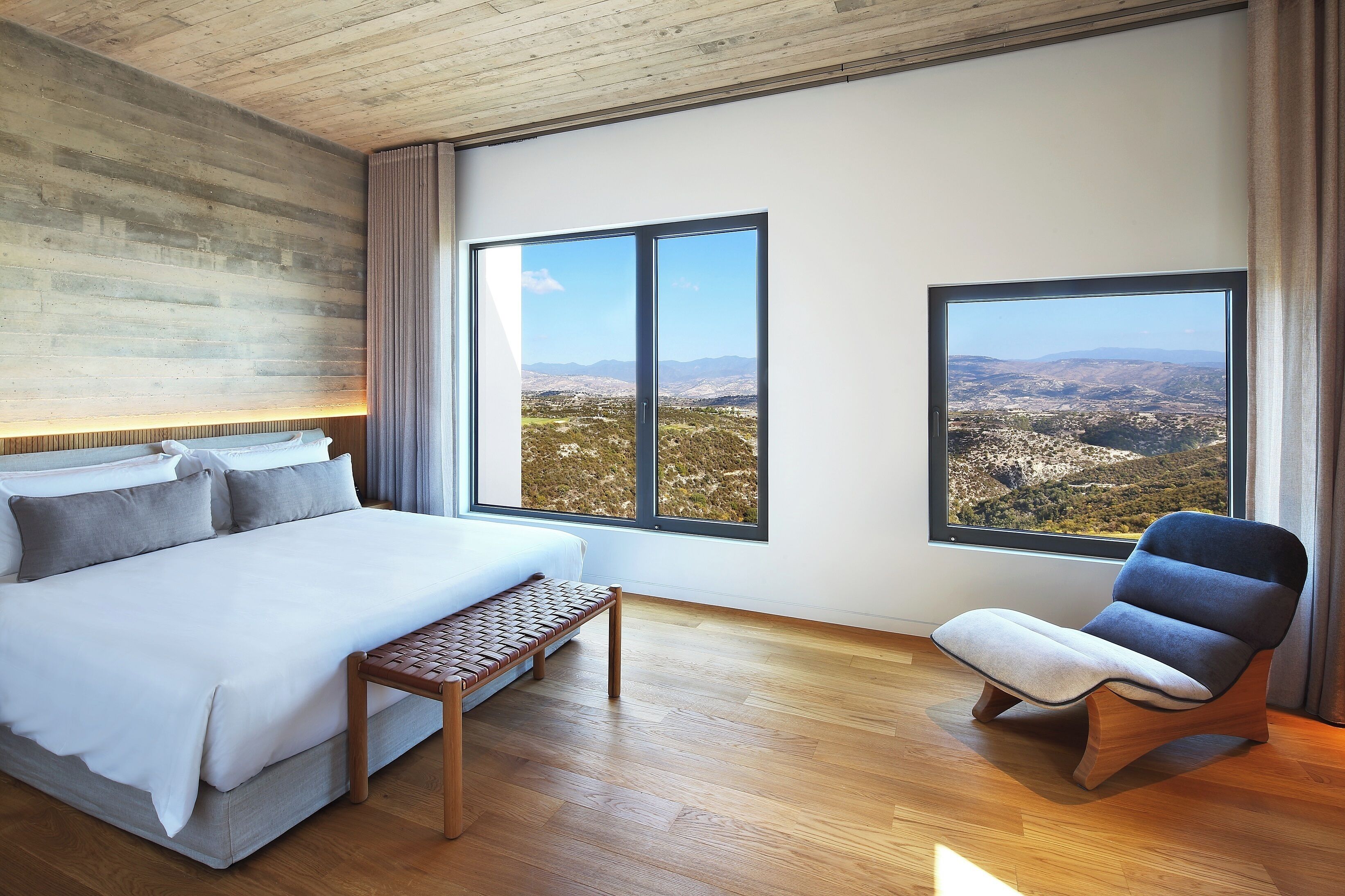 A bedroom with a large bed, a bench, and two windows offering a view of a mountainous landscape.