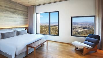 A bedroom with a large bed, a bench, and two windows offering a view of a mountainous landscape.