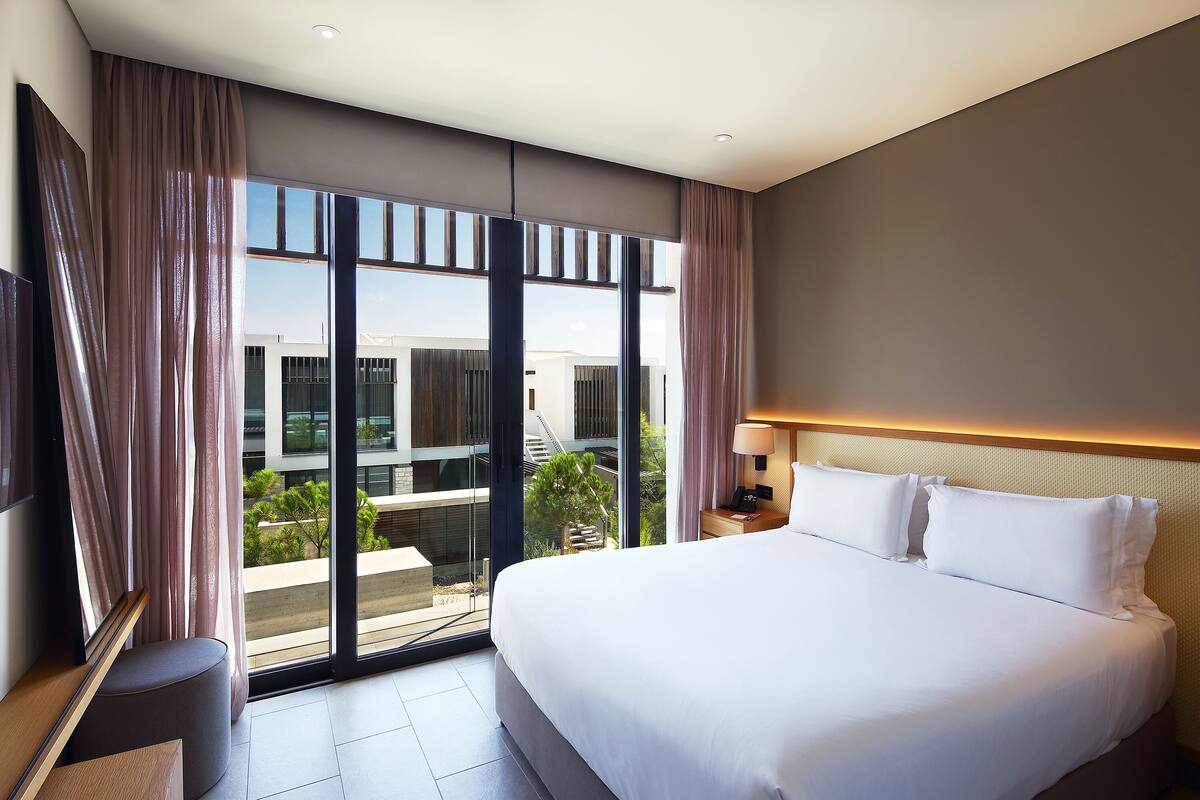 A modern hotel room with a large bed, a TV, and a balcony with a view of the city.