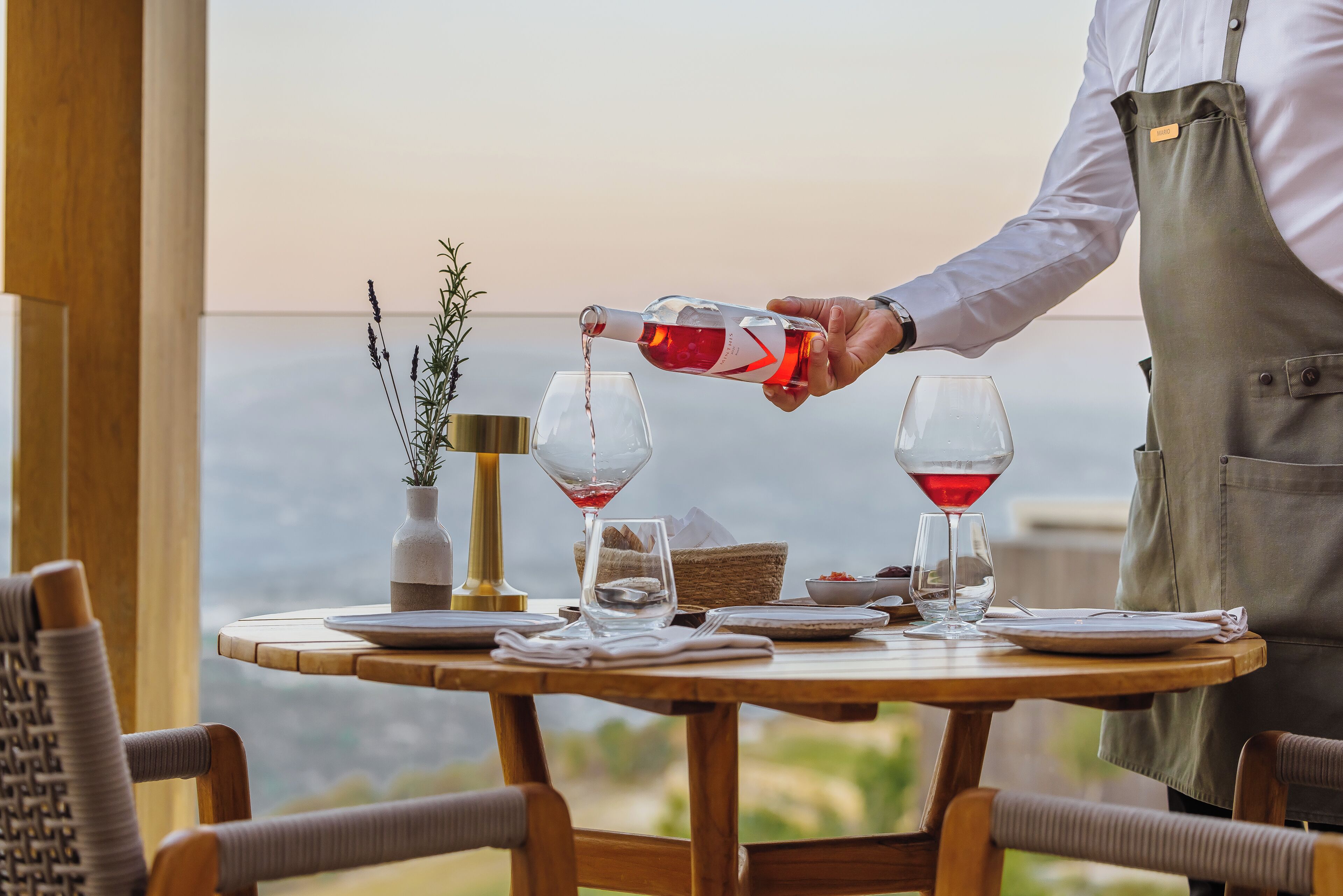 A table with two wine glasses, a vase with plants, and a bottle being poured into one glass.