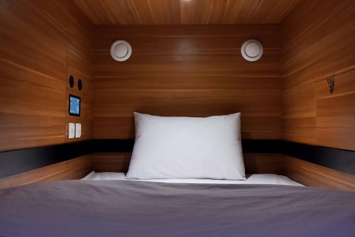 A wooden-paneled room with a bed, two wall-mounted lights, and a small control panel.