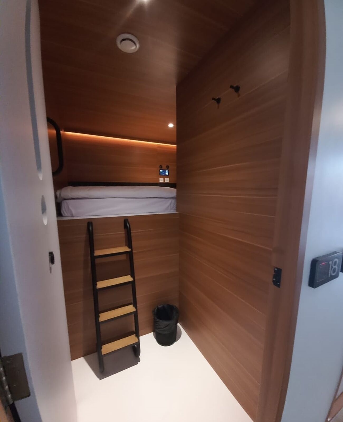A compact room with a bunk bed, a ladder, a trash bin, and a digital display on the wall.