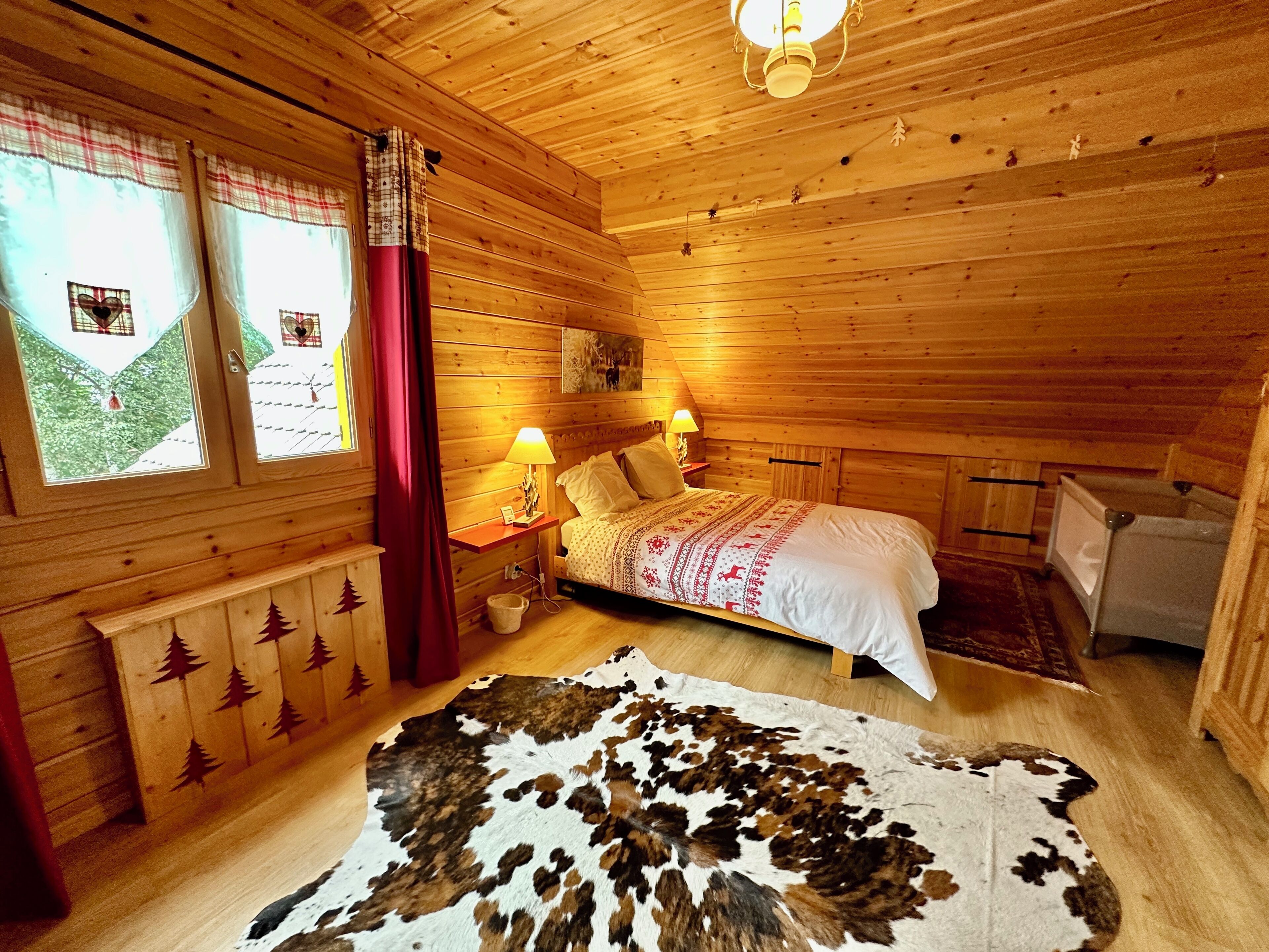 Un dormitorio rústico de cabaña de madera con una cama, dos lámparas de noche, un ventanal con cortinas y un aparador de madera con detalles de hojas.