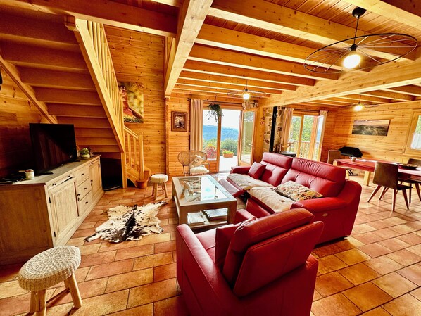Interior acolhedor de uma cabana de madeira, com um sofá vermelho, uma mesa de jantar de madeira e uma televisão sobre um armário.