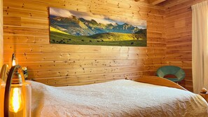 A wooden cabin room with a bed, a painting of a mountain landscape, a chair, and a lamp.
